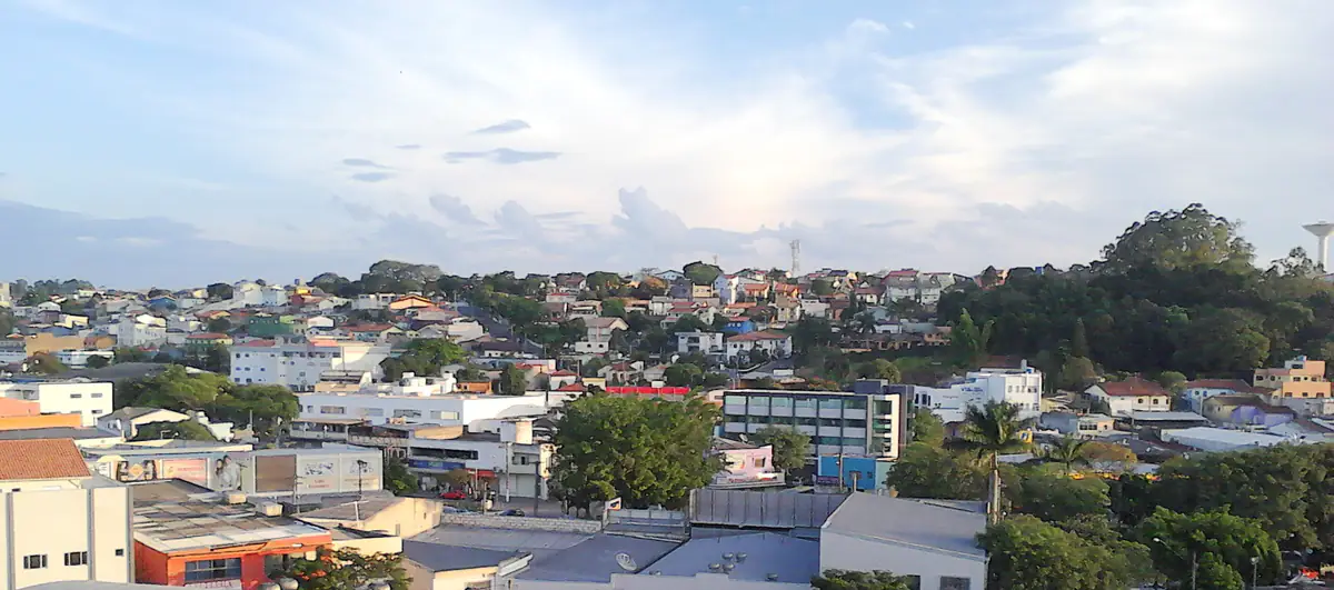 Vista do centro de Arujá (SP) do alto, com casas no morro, árvores e céu azul com nuvens altas no fim da tarde