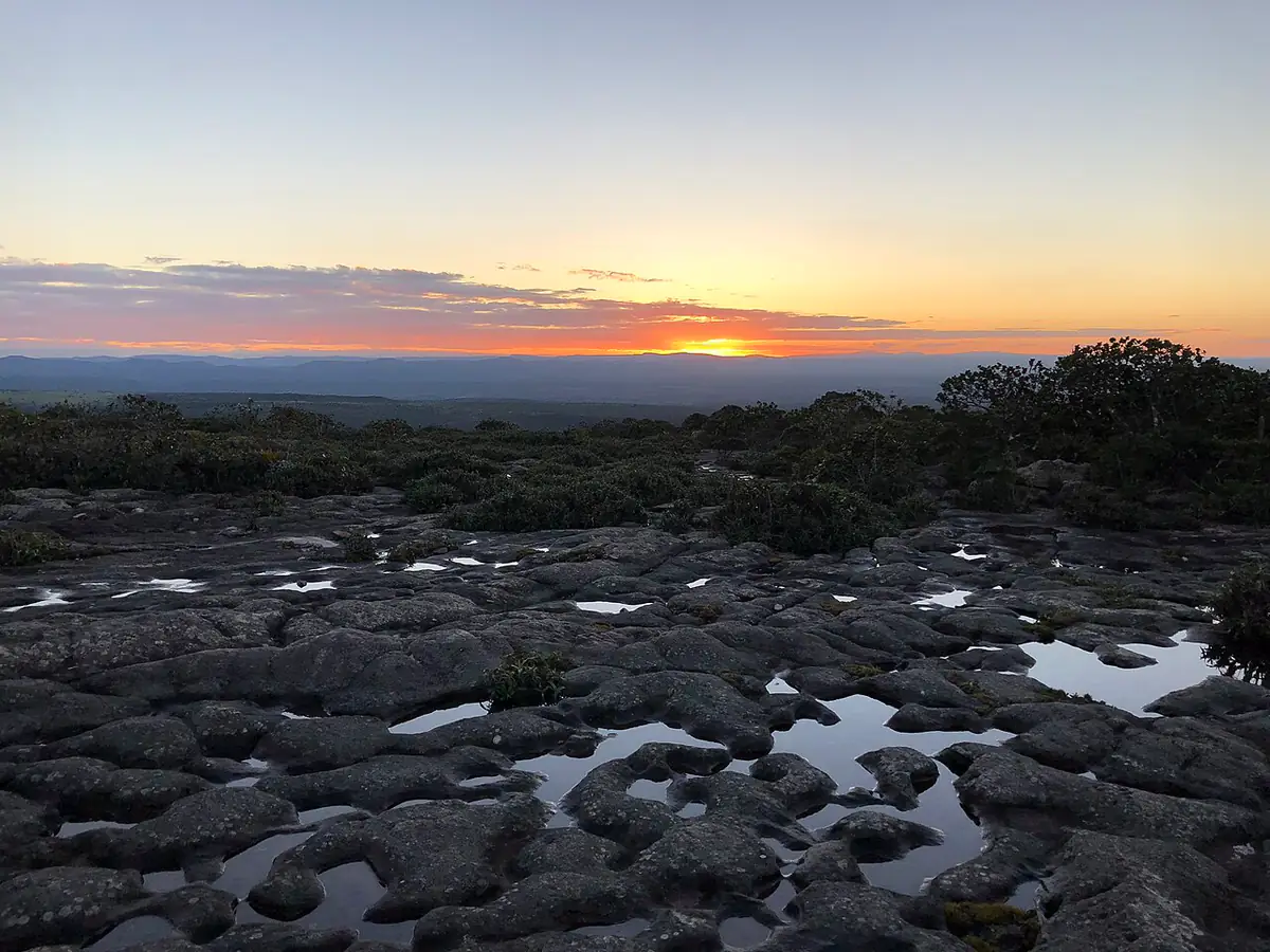 Pôr do sol sobre a serra, visto do alto de uma rocha escura com poças de água da chuva e arbustos baixos