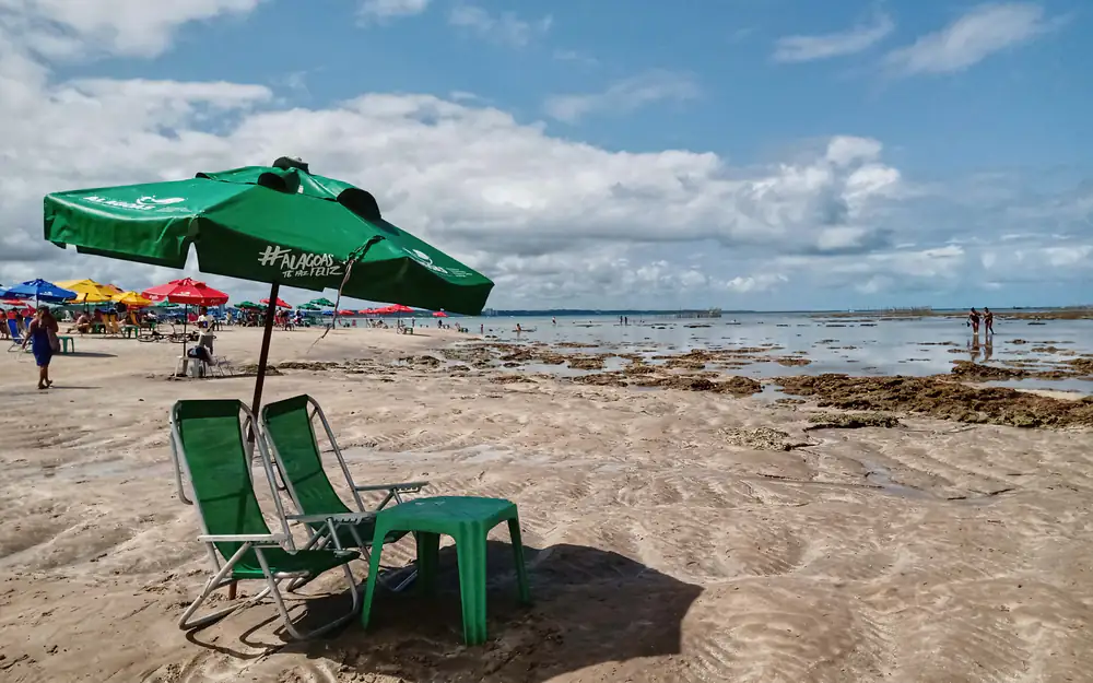 Praia no litoral brasileiro com guarda-sol verde, cadeiras e céu com nuvens de verão