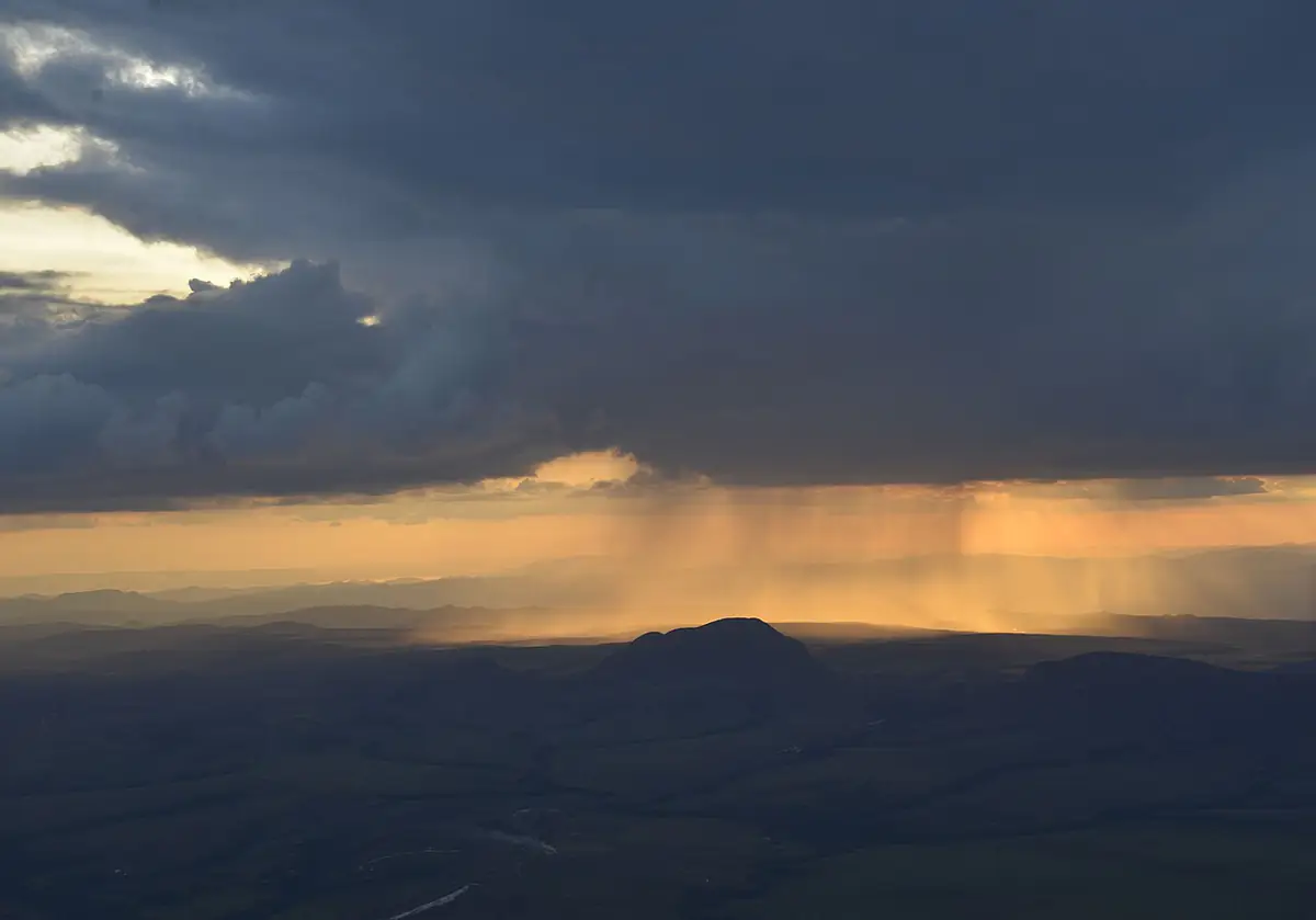 Chuva caindo sob nuvens escuras sobre os morros da Chapada dos Veadeiros, com luz alaranjada ao fundo