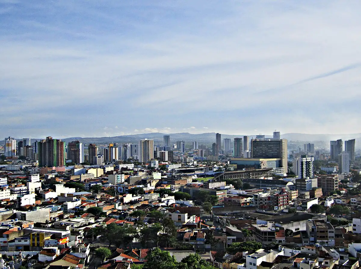 Prédios da cidade de Caruaru vistos do alto, com morros ao fundo e céu azul com nuvens finas