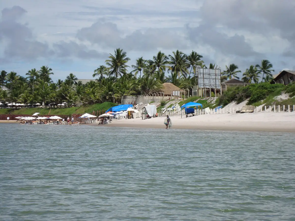 Praia de Porto de Galinhas com mar calmo em primeiro plano, areia clara, coqueiral ao fundo e céu com nuvens carregadas