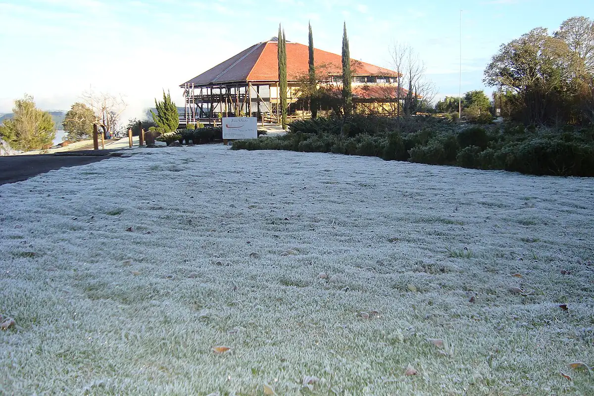 Gramado coberto de geada branca numa manhã de céu limpo, com um pavilhão de telhado vermelho e ciprestes ao fundo
