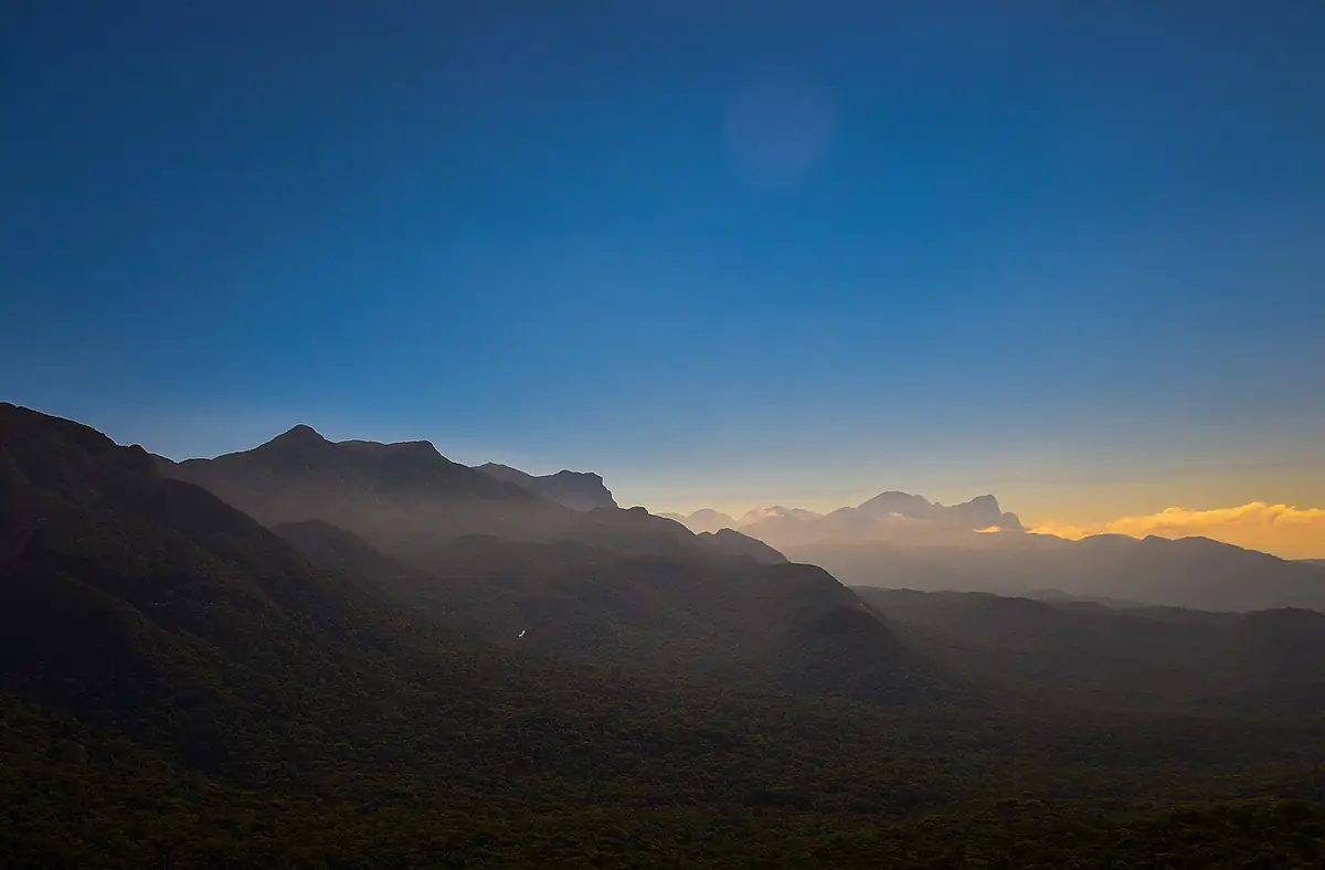 Cristas da Serra do Mar cobertas de mata e de bruma, sob céu azul, com a luz do fim da tarde ao fundo