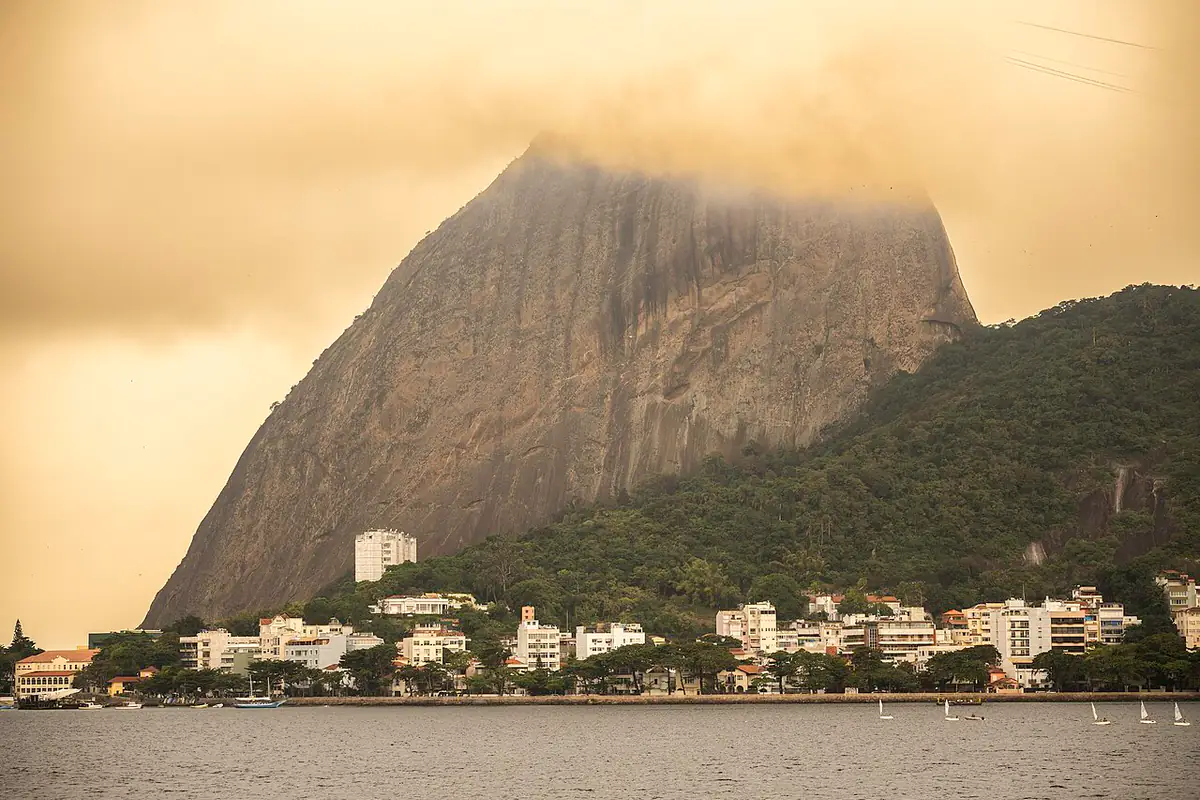 Pão de Açúcar com o topo coberto por nuvens, visto da Baía de Guanabara, com prédios e mata na base do morro