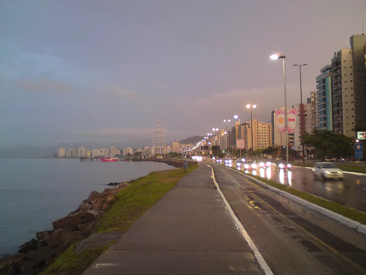 Avenida Beira-Mar de Florianópolis com o asfalto molhado, a baía à esquerda, prédios à direita e céu encoberto ao entardecer