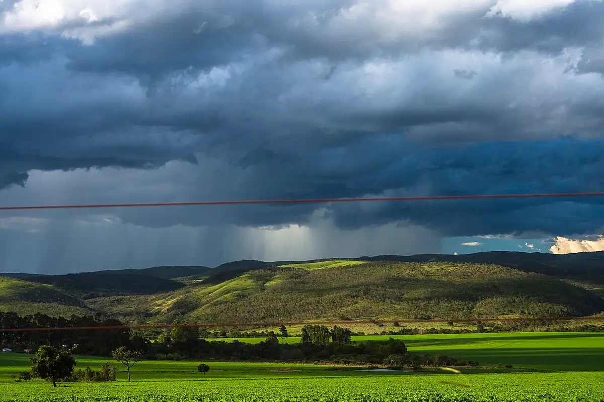 Nuvens escuras de tempestade com chuva caindo ao longe sobre morros e lavoura verde, na estrada entre Alto Paraíso de Goiás e São Jorge