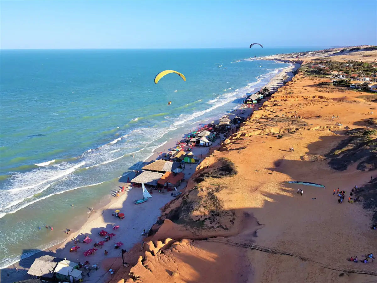 Vista aérea da praia de Canoa Quebrada, em Aracati, com falésias de areia alaranjada, barracas na faixa de areia, ondas e parapentes sobre o mar sob céu azul