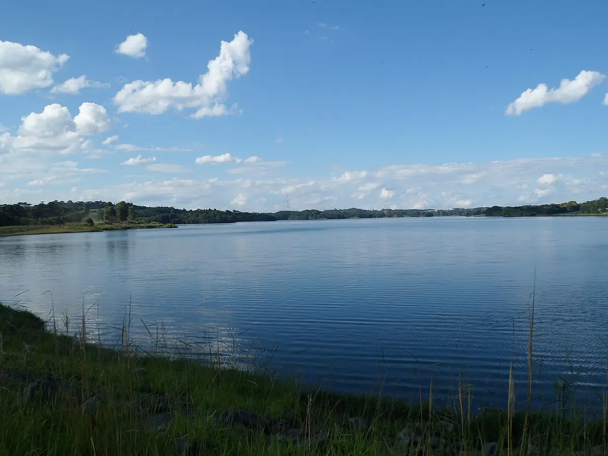Represa do Passaúna em Araucária, com margem de capim, céu azul e nuvens brancas