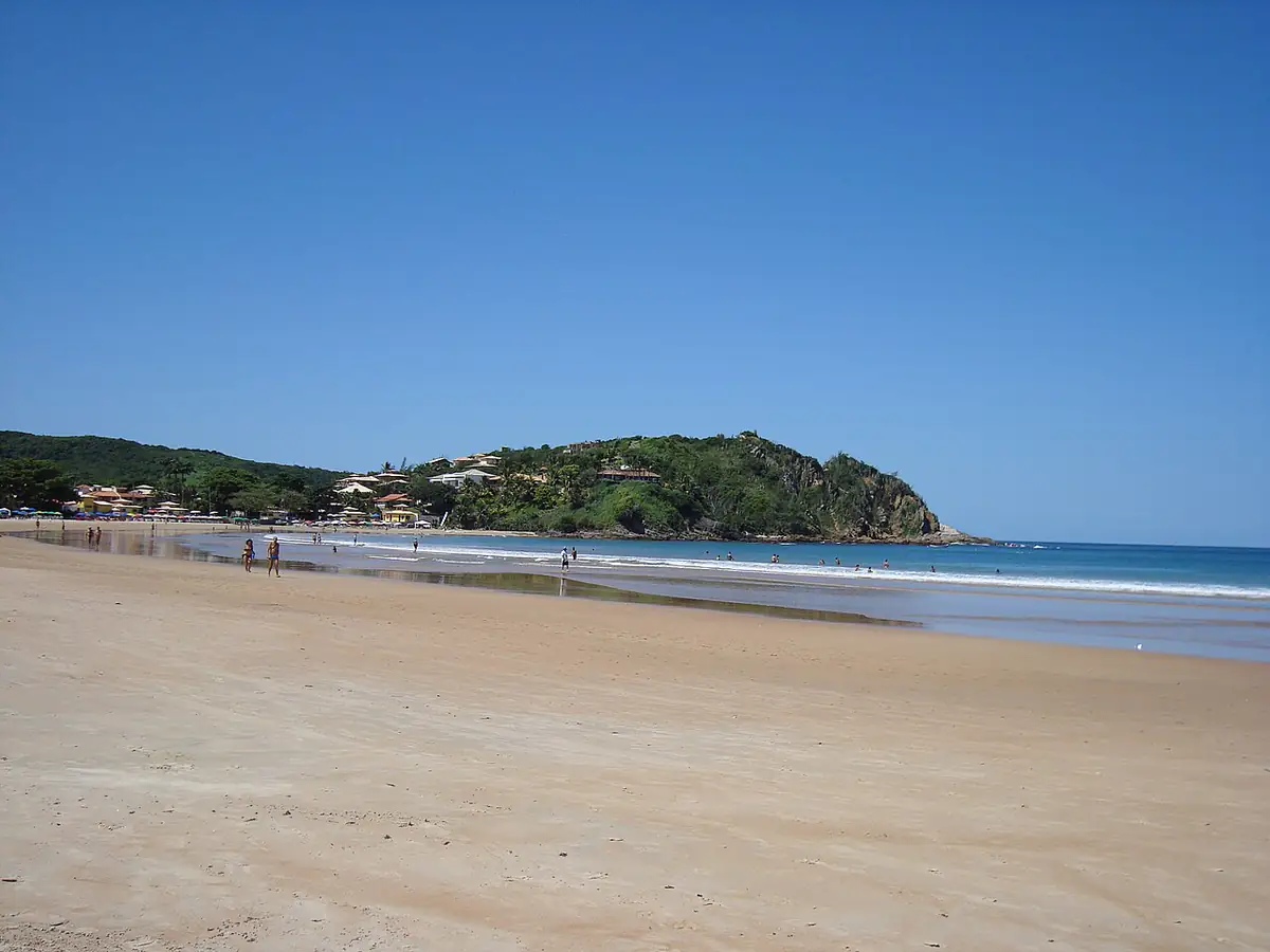 Praia de areia clara em Armação dos Búzios, com morro verde ao fundo e céu azul sem nuvens