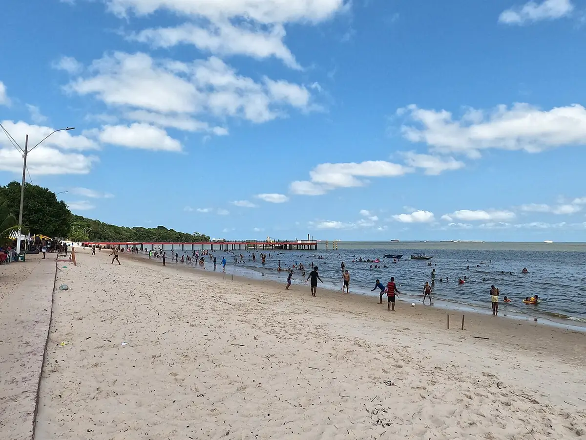 Praia de areia clara à beira do rio, com trapiche ao fundo e nuvens no céu azul