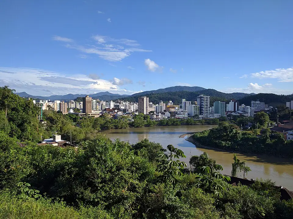 Vista de Blumenau a partir do Morro do Aipim, com prédios do centro, o rio Itajaí-Açu e morros ao fundo sob céu azul com nuvens