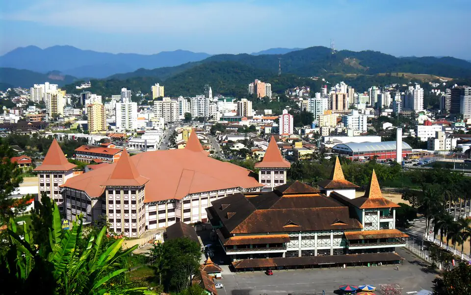 Vista do centro de Brusque com prédios entre colinas verdes e montanhas ao fundo sob céu azul