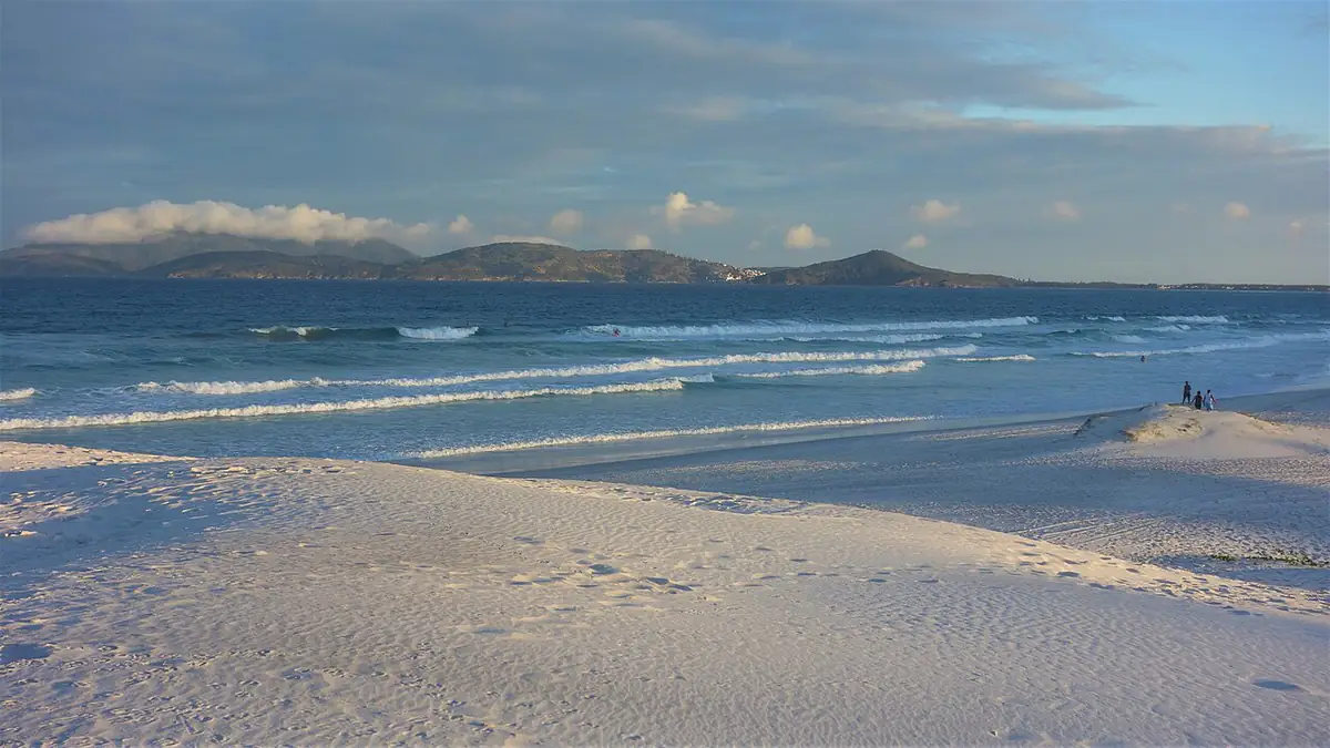 Praia de areia clara em Cabo Frio, com ondas e morros ao fundo sob céu parcialmente nublado
