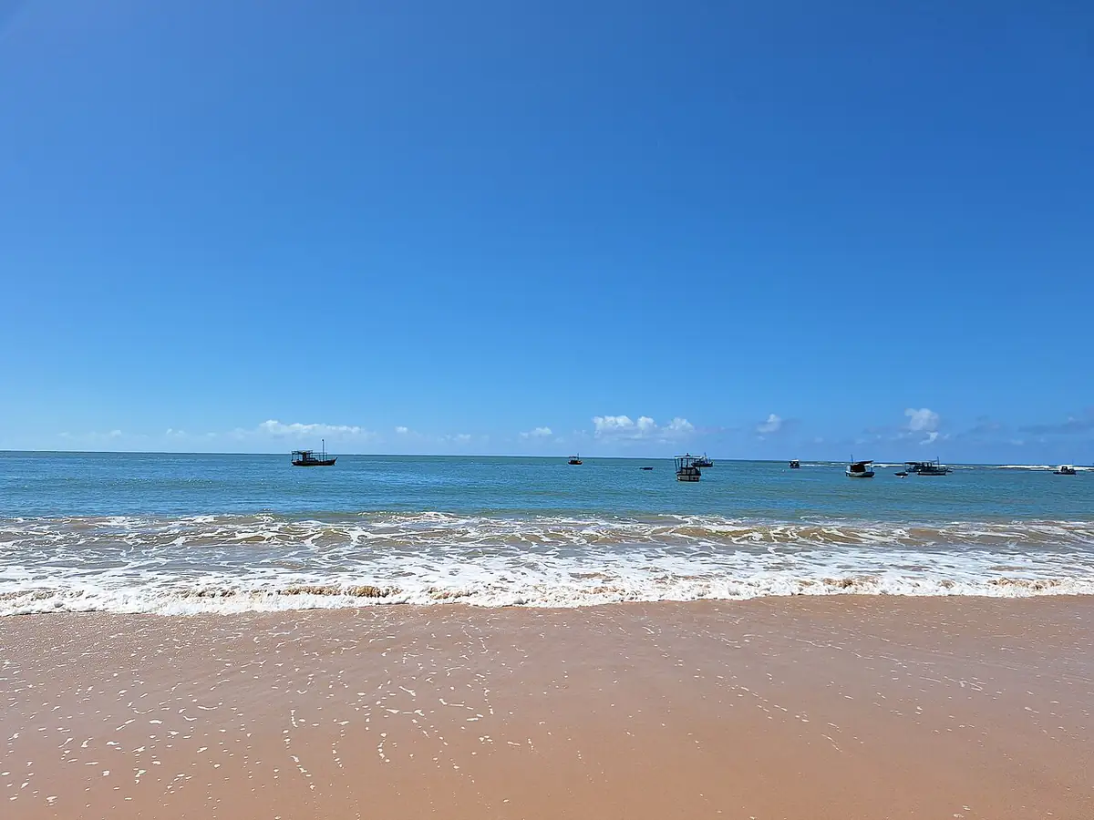 Mar com ondas e barcos de pesca na praia de Itacimirim, em Camaçari, sob céu azul quase sem nuvens
