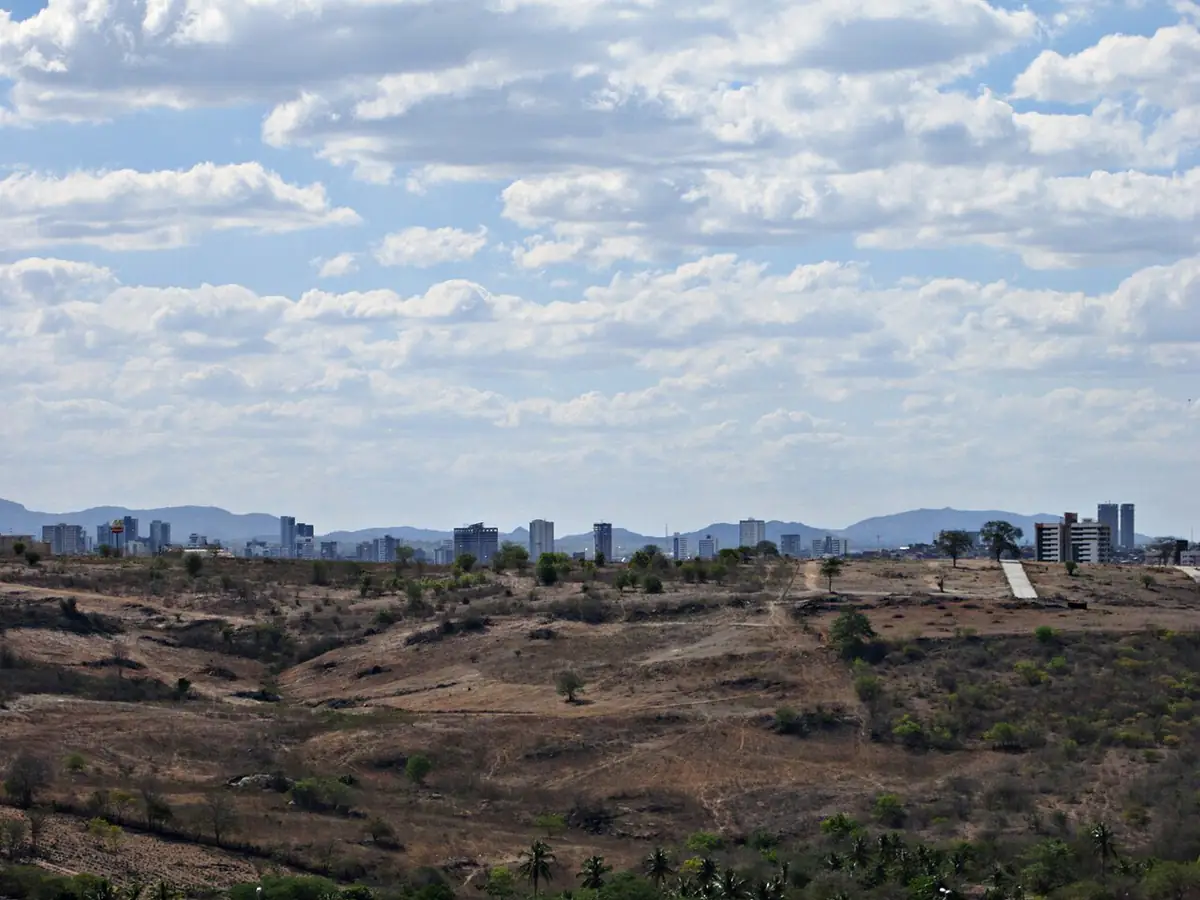 Prédios de Caruaru ao fundo, vistos de uma encosta de vegetação seca, sob céu com muitas nuvens