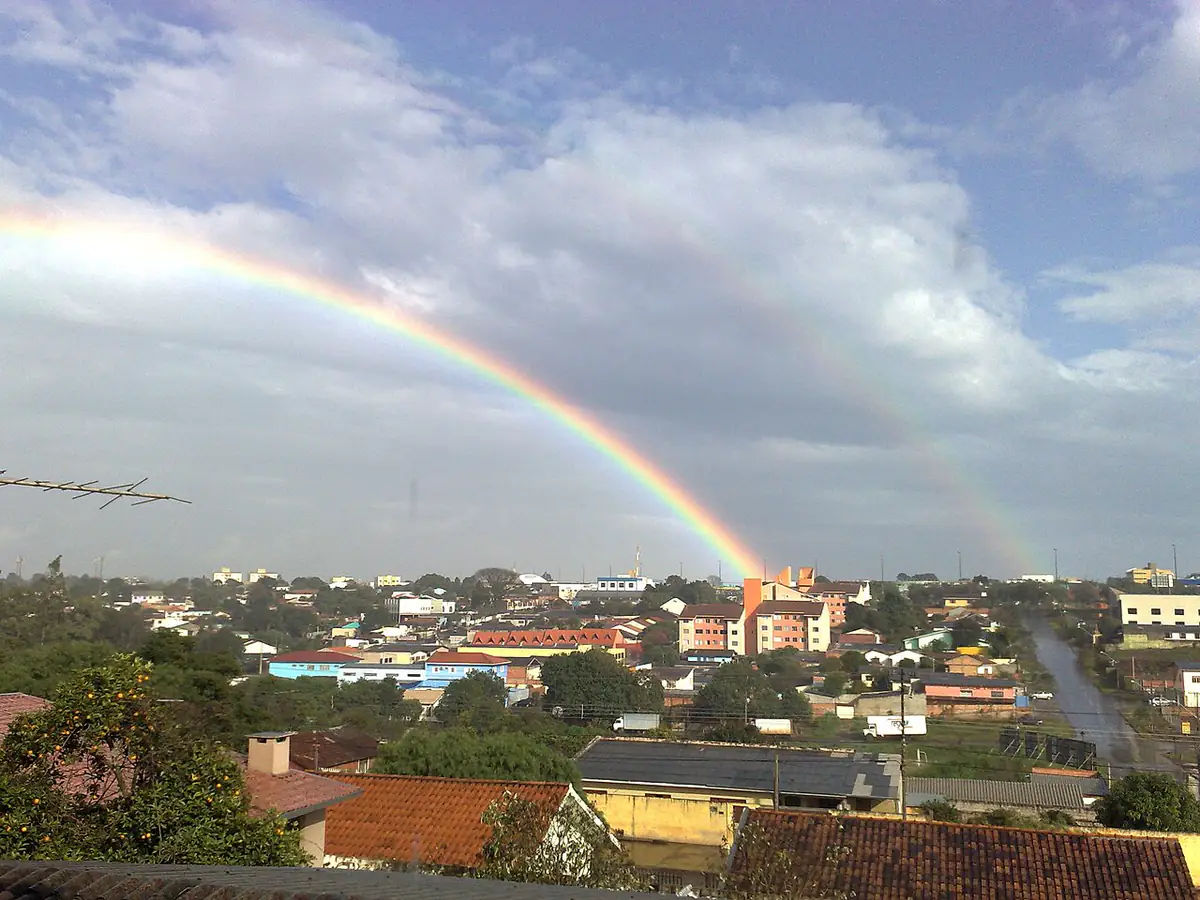 Arco-íris duplo sobre casas e prédios do bairro Neva, em Cascavel, sob nuvens carregadas