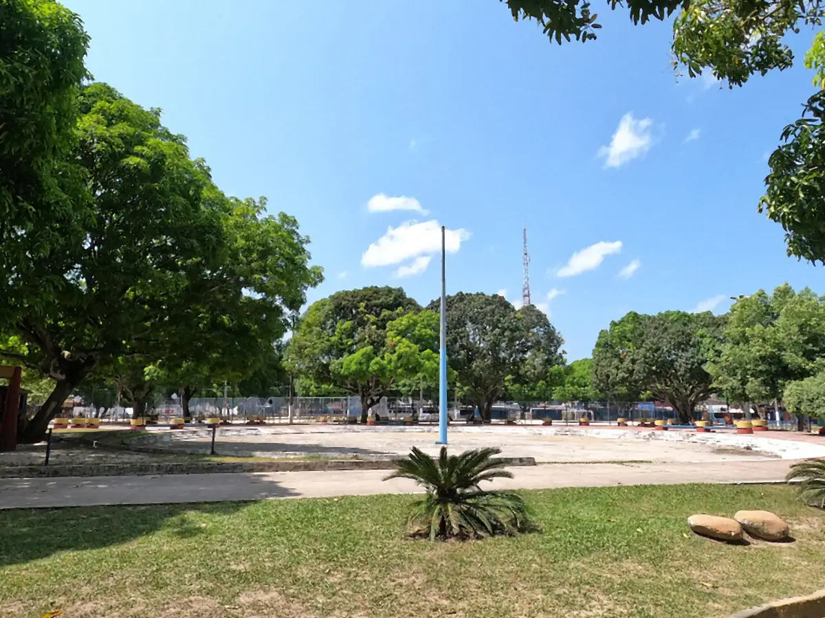Praça arborizada com mangueiras e gramado, sob céu azul com poucas nuvens