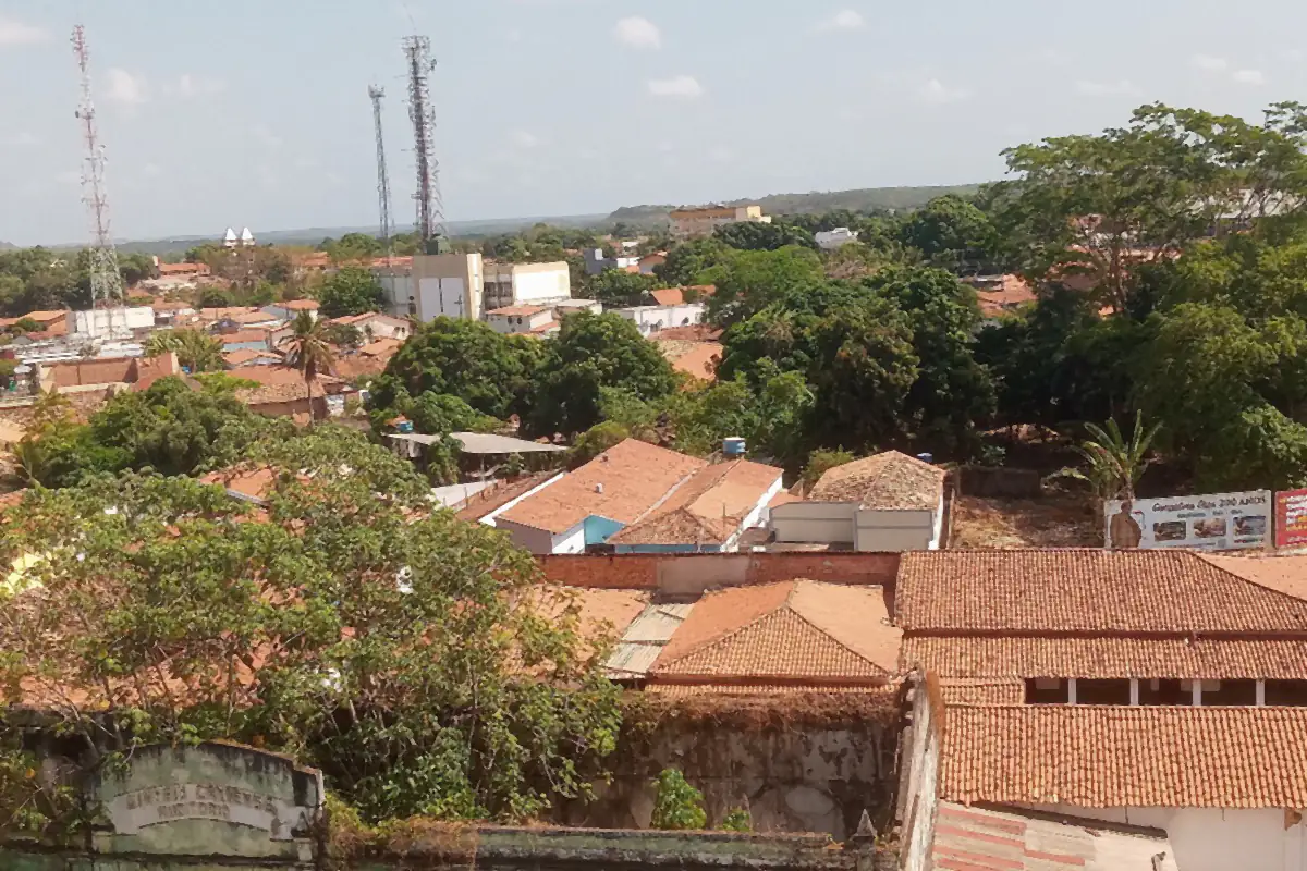 Telhados e árvores de Caxias vistos do alto, com torres de telecomunicação e céu com poucas nuvens
