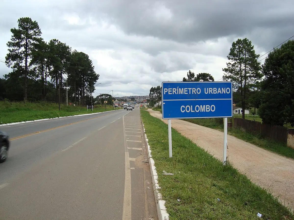 Estrada na entrada do perímetro urbano de Colombo sob céu carregado, com pinheiros ao lado da pista