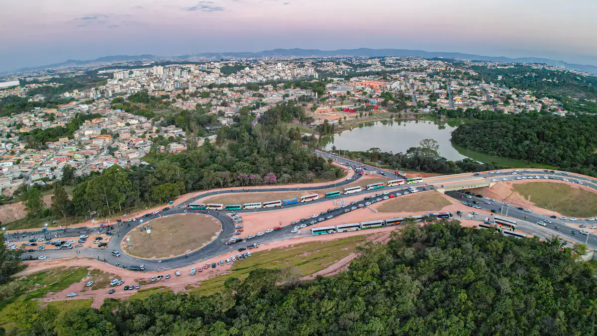 Vista aérea ao entardecer de Contagem (MG), com prédios e bairros residenciais, uma lagoa cercada de vegetação, rodovia com tráfego intenso e serras ao fundo sob um céu com nuvens.