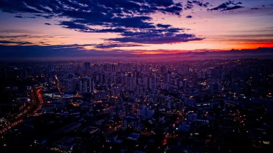 Vista aérea de Criciúma ao anoitecer, com prédios iluminados e céu com nuvens escuras e faixa laranja no horizonte