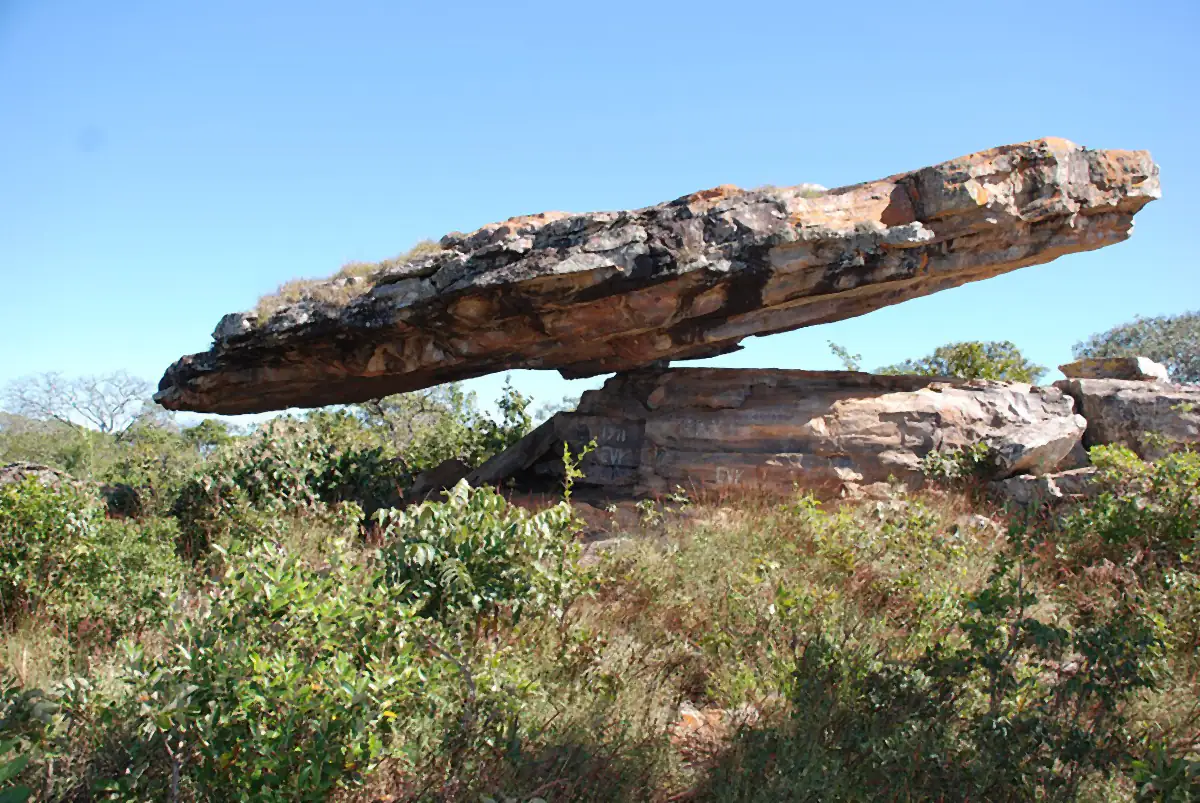 Grande laje de pedra apoiada em outra rocha, em equilíbrio, com cerrado e céu azul ao redor