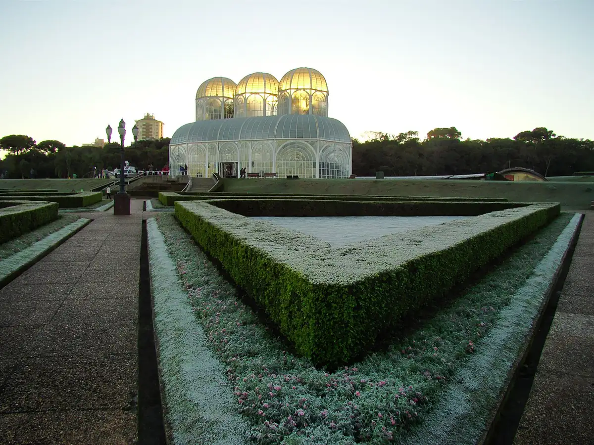 Geada cobrindo os canteiros do Jardim Botânico de Curitiba ao amanhecer, com a estufa de vidro ao fundo