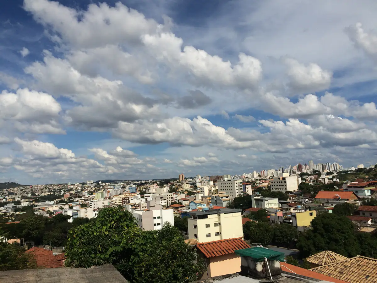 Vista de Divinópolis sob céu azul com nuvens cúmulos, prédios residenciais e telhados em primeiro plano
