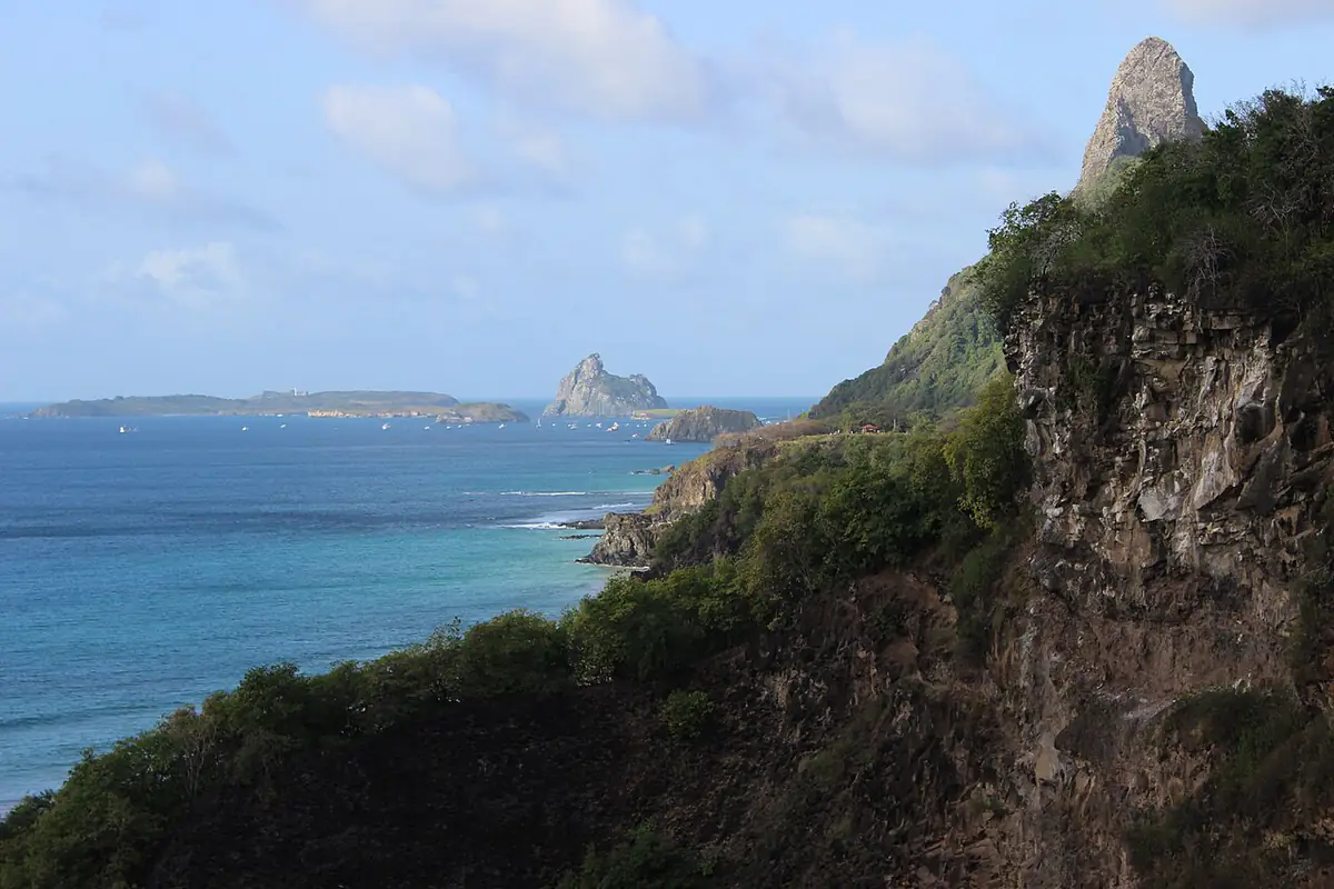 Costa de Fernando de Noronha com falésia verde, o Morro do Pico ao fundo, mar azul e céu com nuvens