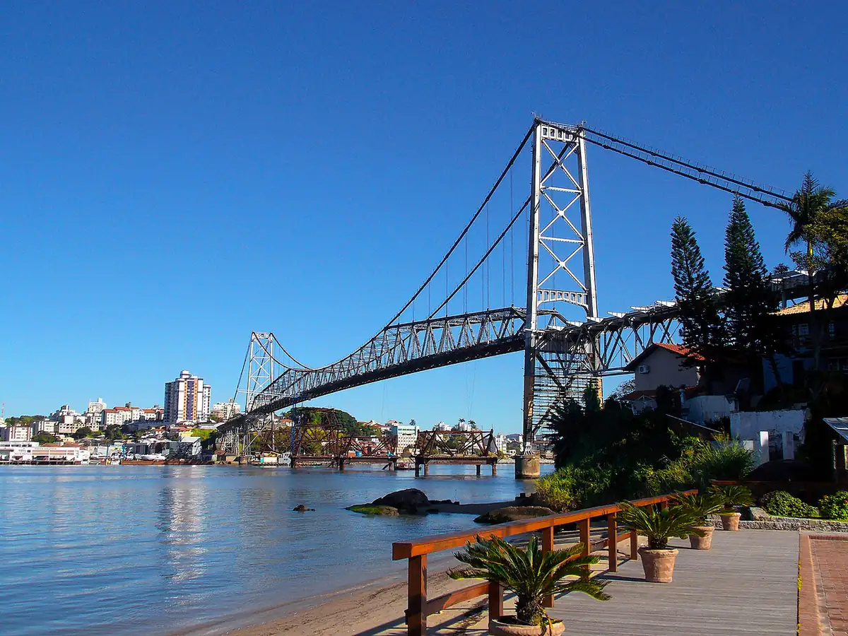 Ponte Hercílio Luz sobre o canal entre a ilha e o continente, em Florianópolis, com céu azul e prédios ao fundo