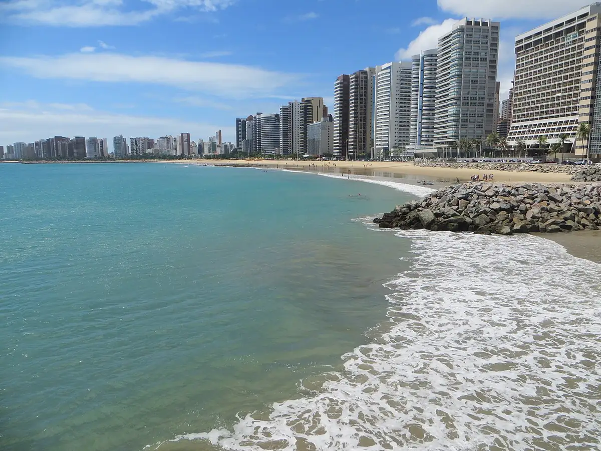 Orla de Fortaleza com mar de cor esverdeada, praia e prédios ao fundo sob céu azul com nuvens