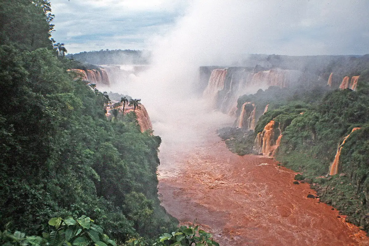 Cataratas do Iguaçu com o rio cheio e de cor avermelhada, entre a mata, com névoa de água no ar