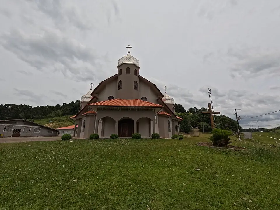 Igreja ucraniana de torres com cúpulas, cercada por gramado e morros com árvores, sob céu nublado, em General Carneiro