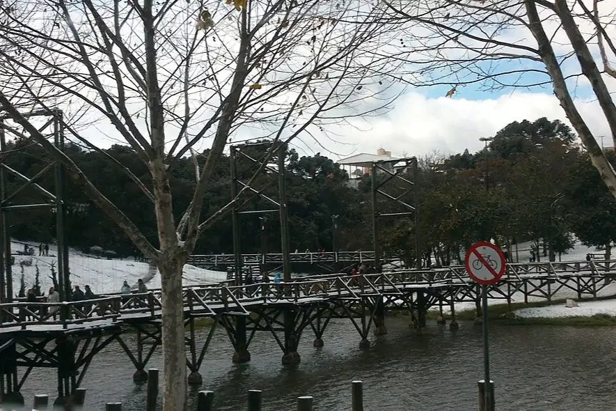 Passarela de madeira sobre um lago, com neve no chão e nas margens e árvores sem folhas, em Guarapuava