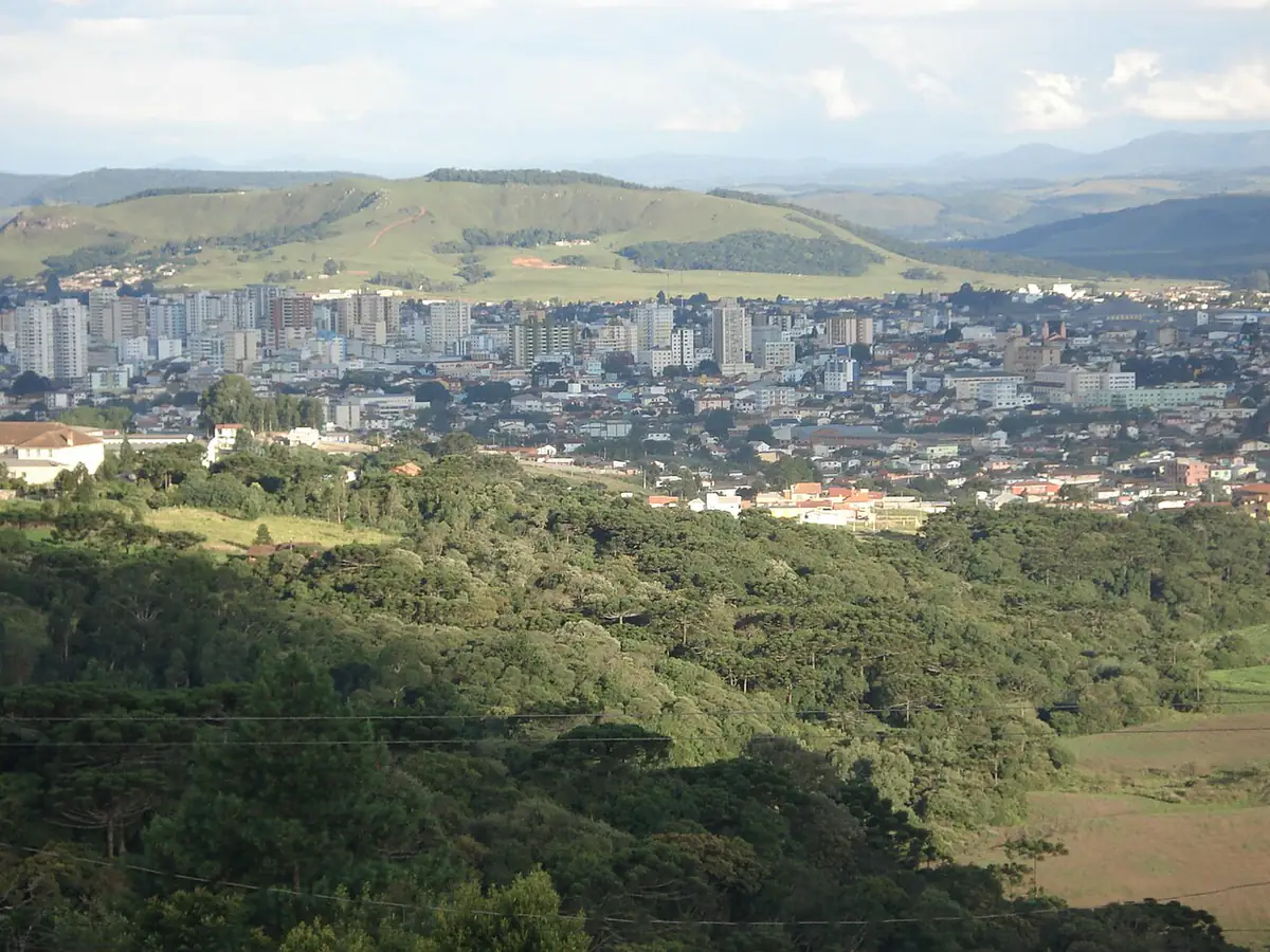 Vista do centro de Lages, com prédios entre colinas de campo e matas de araucária sob céu com nuvens