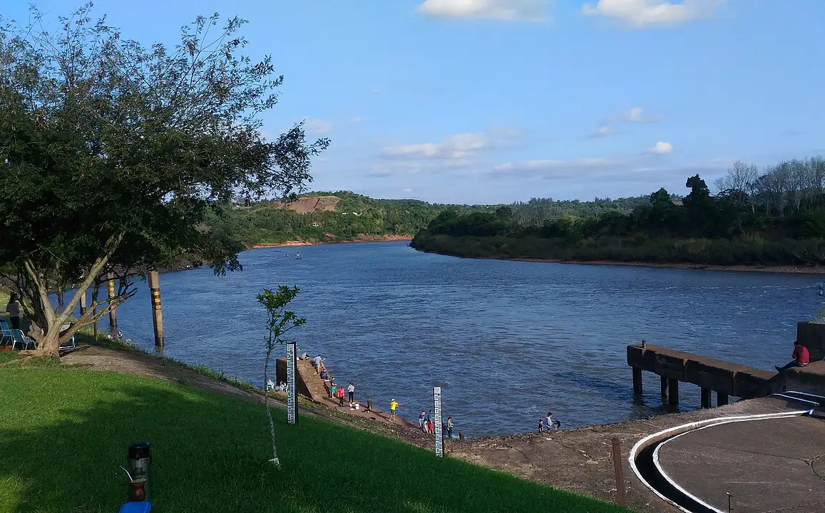 Rio Taquari largo e calmo visto da margem em Lajeado, com réguas de medição do nível da água e mata na outra margem