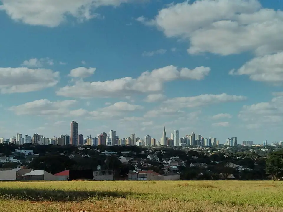 Prédios do centro de Maringá ao fundo, atrás de um terreno de grama, sob céu azul com nuvens