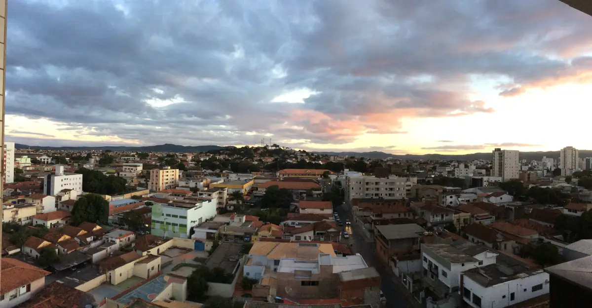 Vista aérea do pôr do sol sobre bairros de Montes Claros, com telhados de barro, prédios e morros arredondados no horizonte.