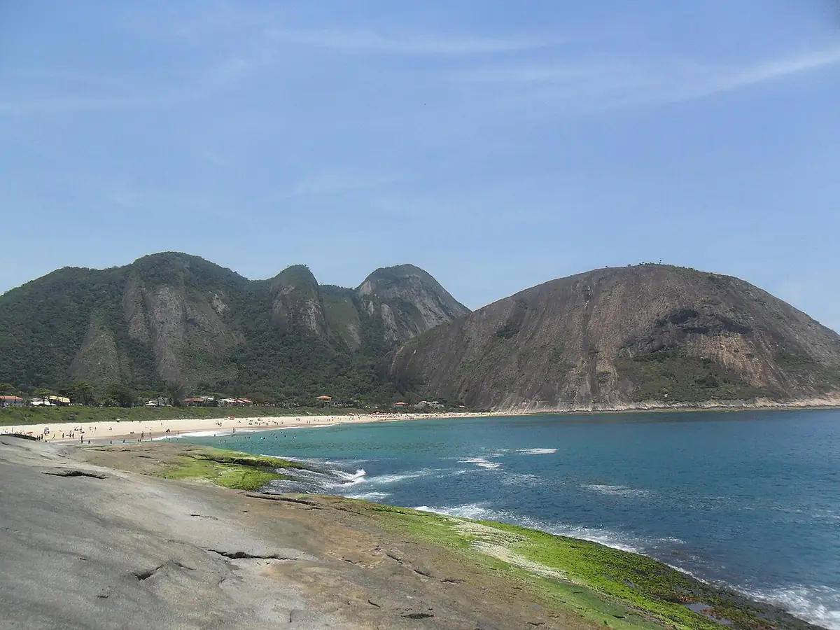 Praia de Itacoatiara, em Niterói, vista de uma laje de pedra, com morros cobertos de mata ao fundo e céu azul