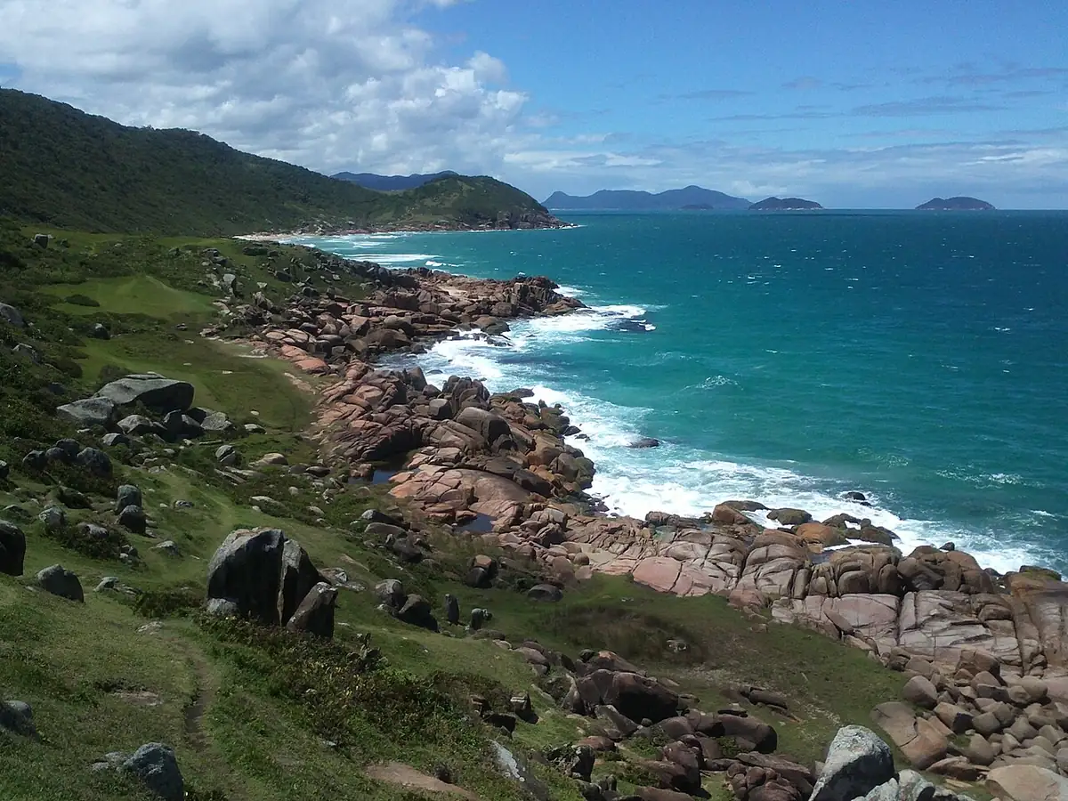 Costa rochosa da praia da Guarda do Embaú, em Palhoça, com ondas, mar verde e uma ilha ao fundo sob céu parcialmente nublado