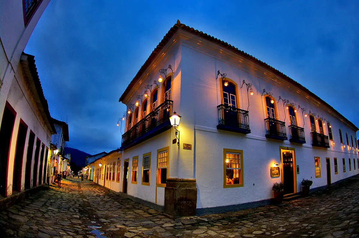 Casarão colonial branco de esquina com lampiões acesos, rua de pedras molhadas e céu azul escuro de fim de tarde no centro histórico de Paraty
