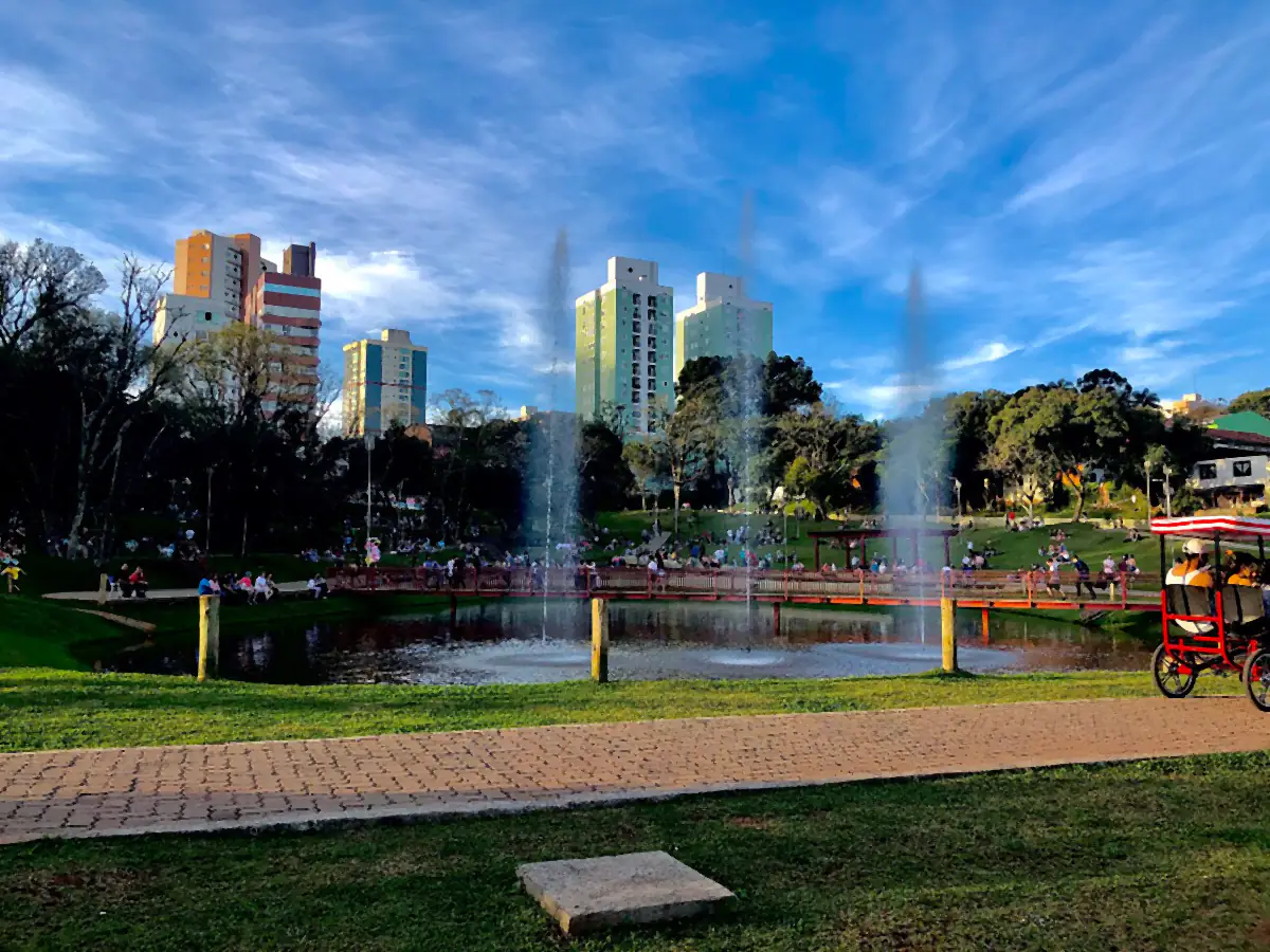 Lago com chafariz no Parque da Gare, em Passo Fundo, com prédios ao fundo e céu azul com nuvens finas