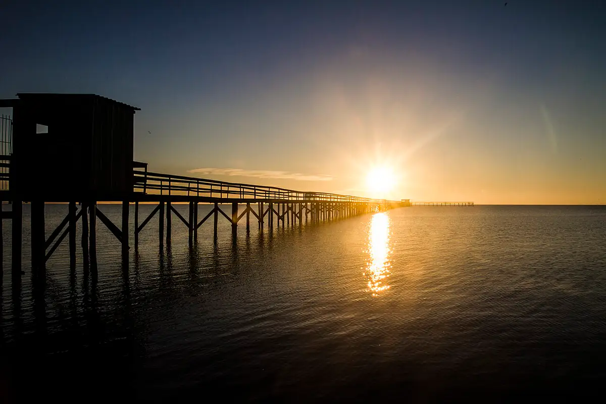 Amanhecer sobre a Lagoa dos Patos, com um longo píer de madeira e o sol baixo refletido na água, na Praia do Laranjal