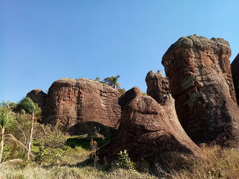 Formações de arenito avermelhado sob céu azul limpo, com vegetação de campo na base, no Parque Estadual de Vila Velha