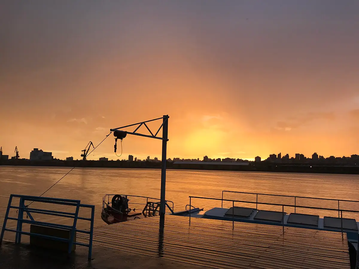 Pôr do sol alaranjado sobre o Lago Guaíba depois da chuva, com guindastes do porto e prédios de Porto Alegre ao fundo