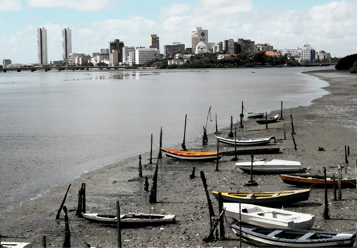 Barcos na maré baixa do Rio Capibaribe, com os prédios do Recife ao fundo sob céu com nuvens