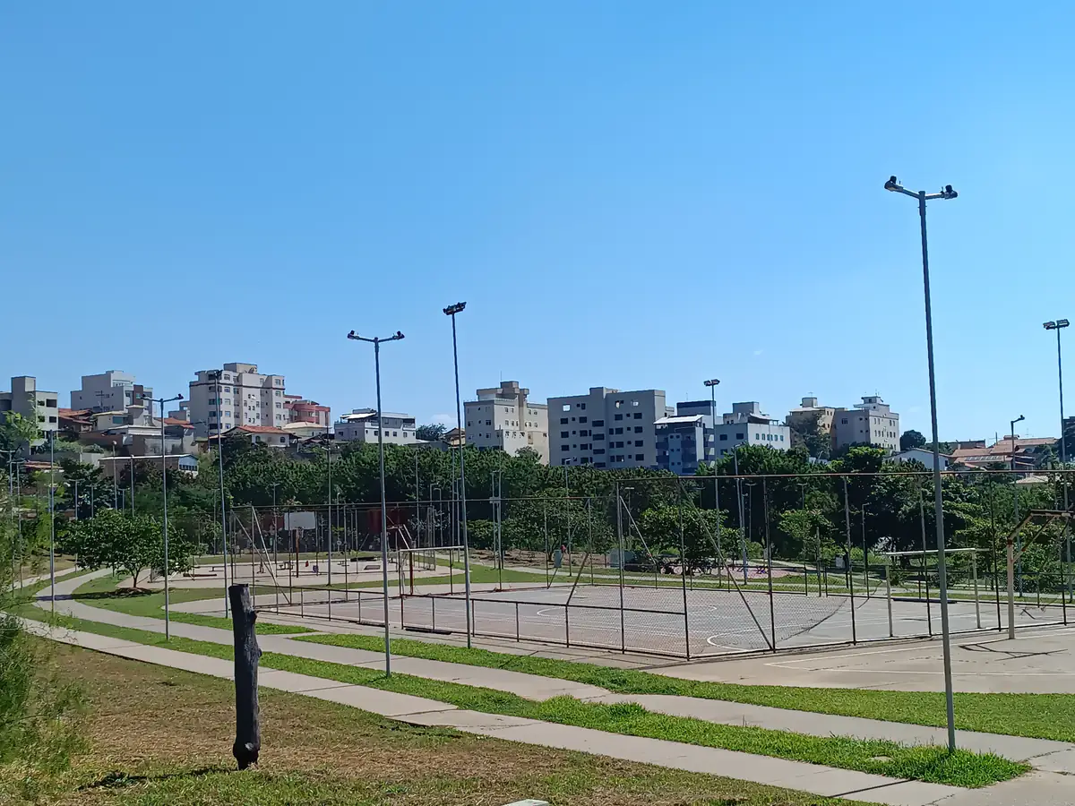 Vista do Parque Ecológico de Ribeirão das Neves, com quadras poliesportivas em primeiro plano, árvores e prédios residenciais ao fundo, sob céu azul sem nuvens