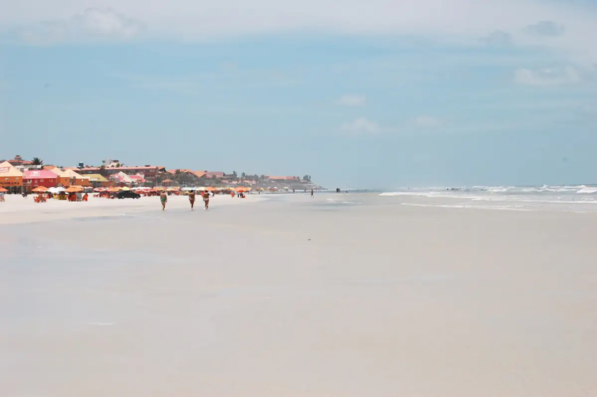 Praia do Atalaia com faixa larga de areia úmida, quiosques ao fundo e céu com nuvens