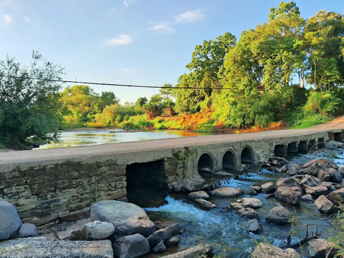 Rio Pardinho correndo entre pedras sob uma ponte baixa de concreto, com mata e céu azul ao fundo